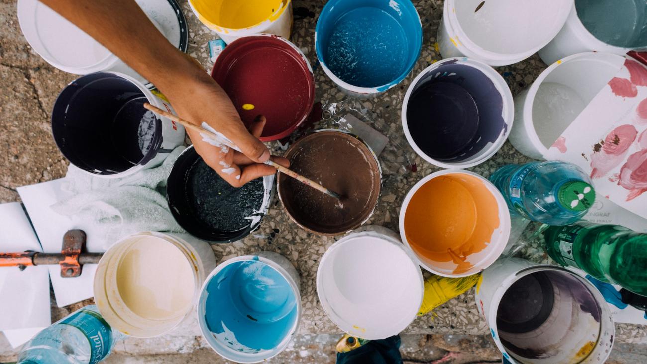 Close-up of a person dipping their paintbrush in a can of paint, among many.