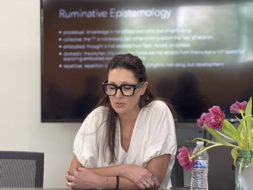Stacey Moran speaks, seated at a table during an event. A slide on a screen behind her says "ruminative epistemology."